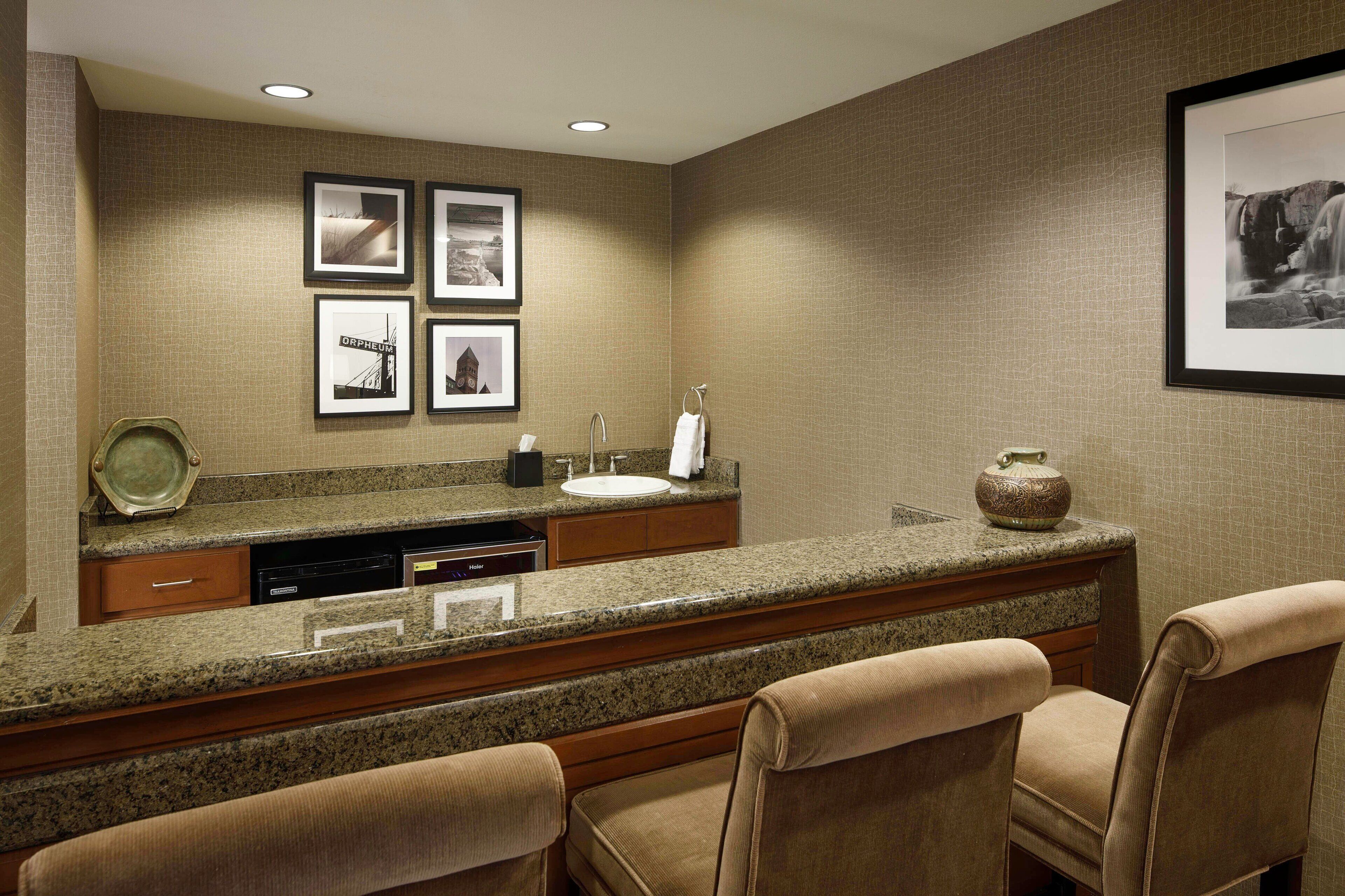 A hotel bathroom with a granite countertop, sink, and two chairs. There are framed pictures on the wall and a vase on the countertop.