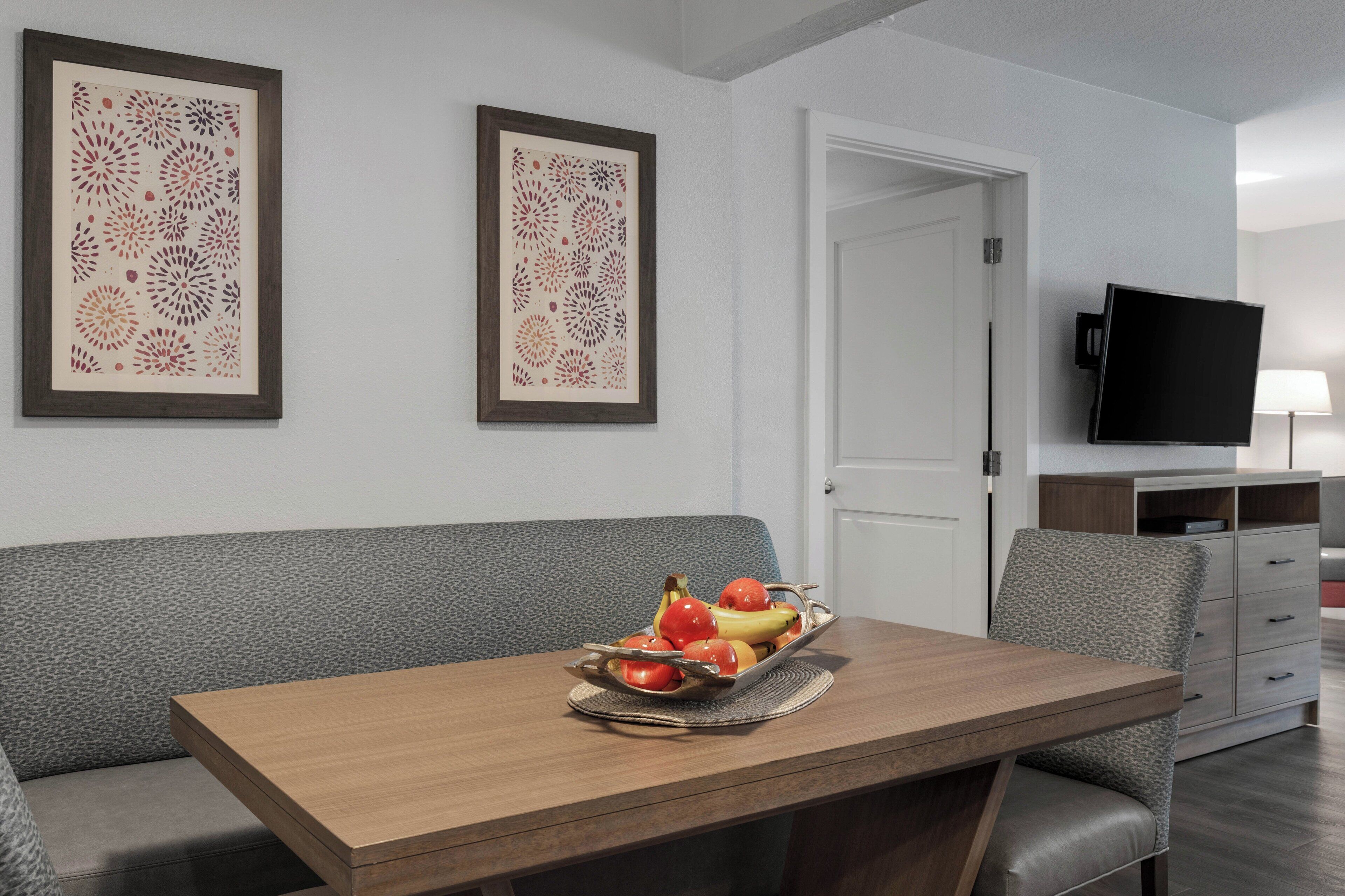 Una sala de estar moderna con una mesa de comedor de madera, un sofá gris y un cuenco de frutas. Hay dos obras de arte enmarcadas en la pared y un televisor a la derecha.