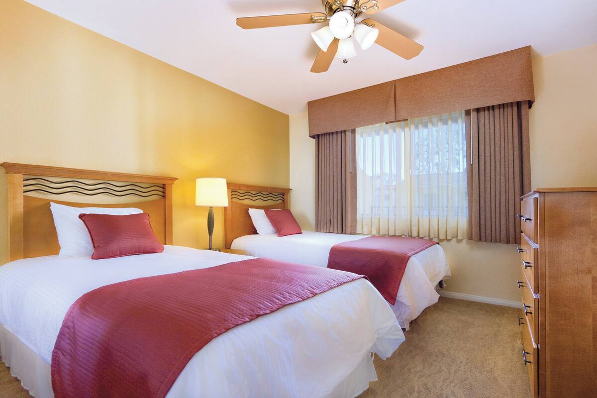 A hotel room with two beds, a ceiling fan, a nightstand with a lamp, a dresser, and a window with curtains.