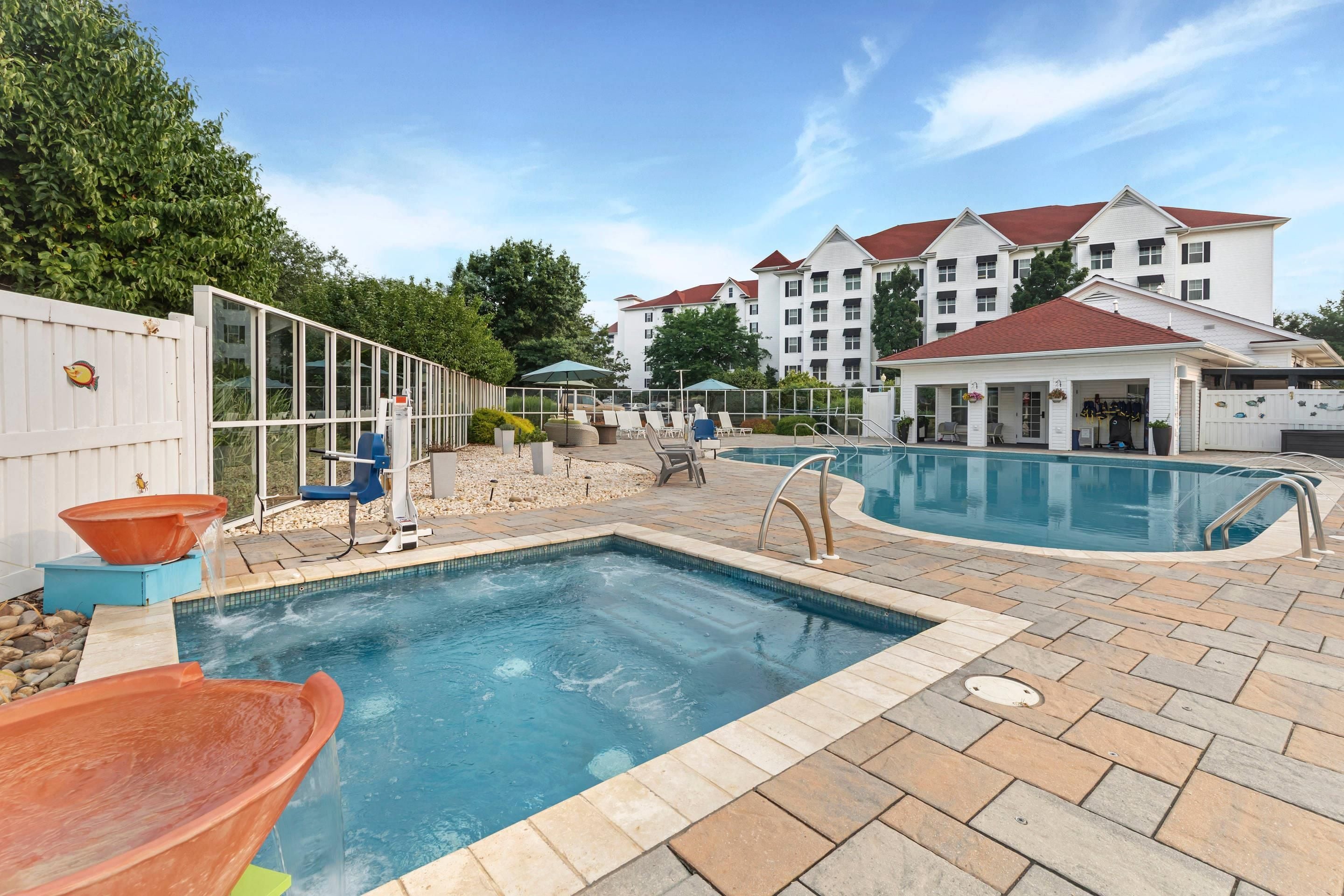 A swimming pool area with a hot tub, lounge chairs, and a building with a red roof in the background.