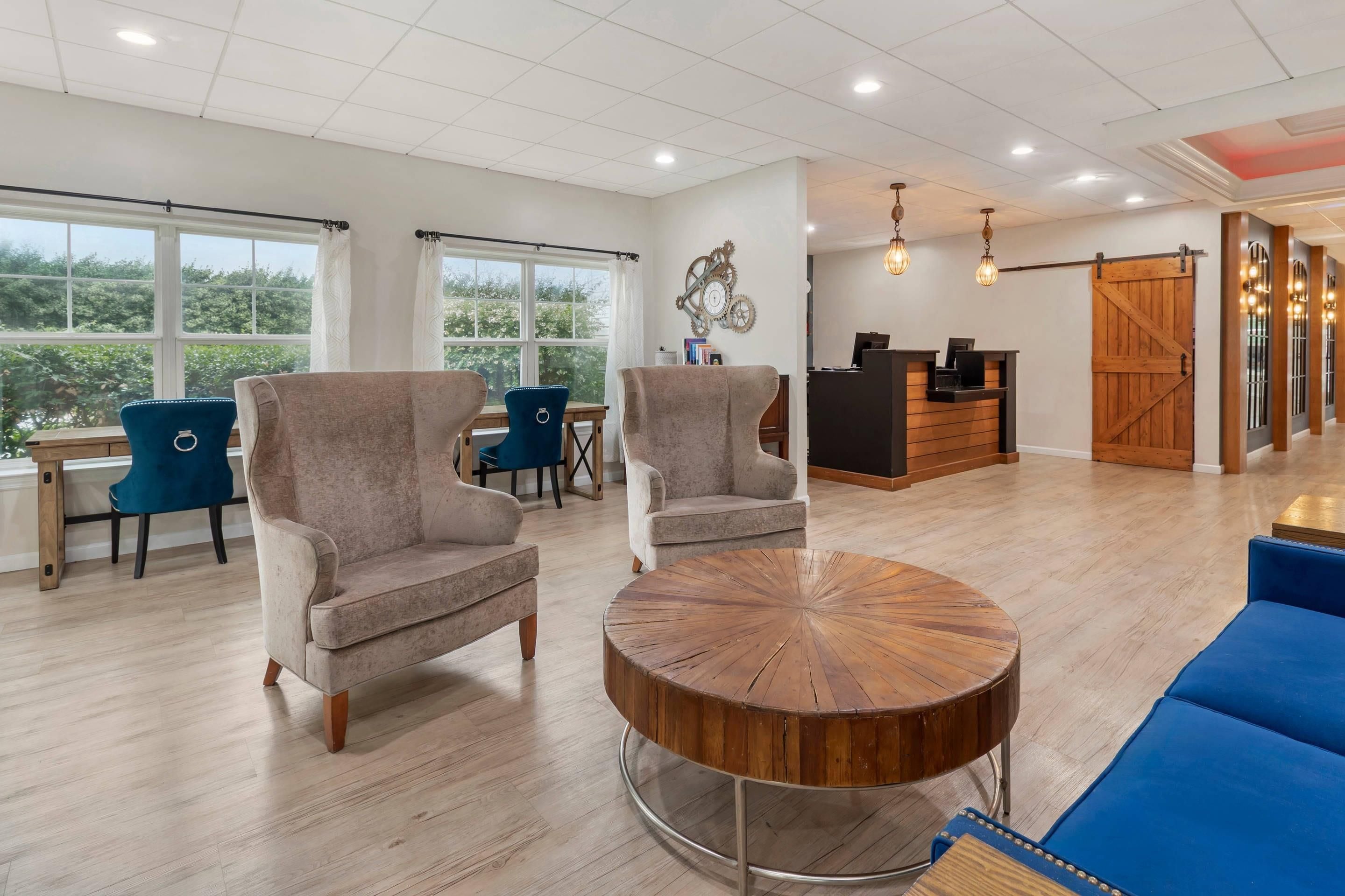 A spacious room with a reception desk, seating area with armchairs and a round coffee table, and large windows offering a view of greenery.