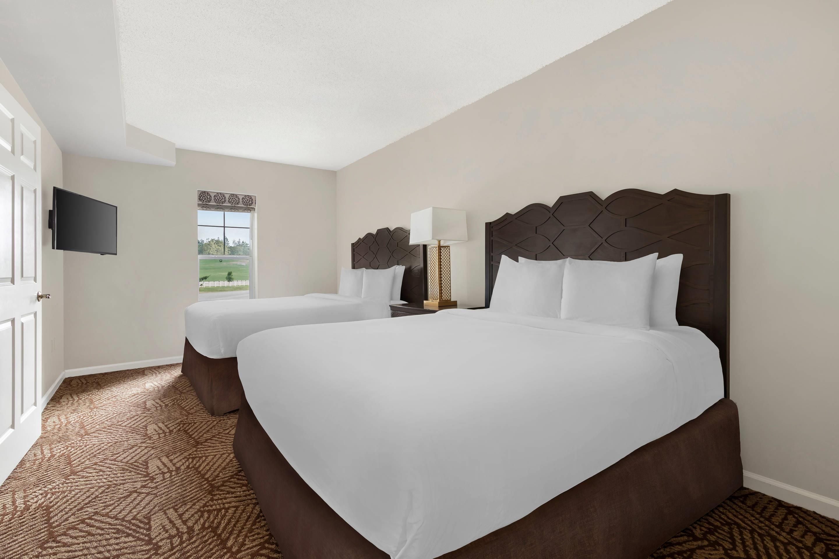 A hotel room with two beds, a TV mounted on the wall, and a window with a view of the outdoors.