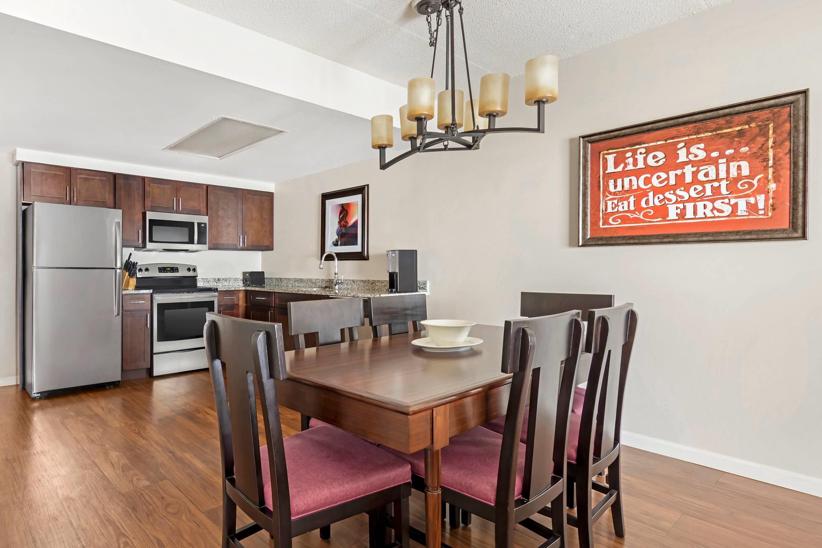 A modern kitchen with stainless steel appliances, a dining area with wooden chairs and a table, and a wall with a motivational quote.