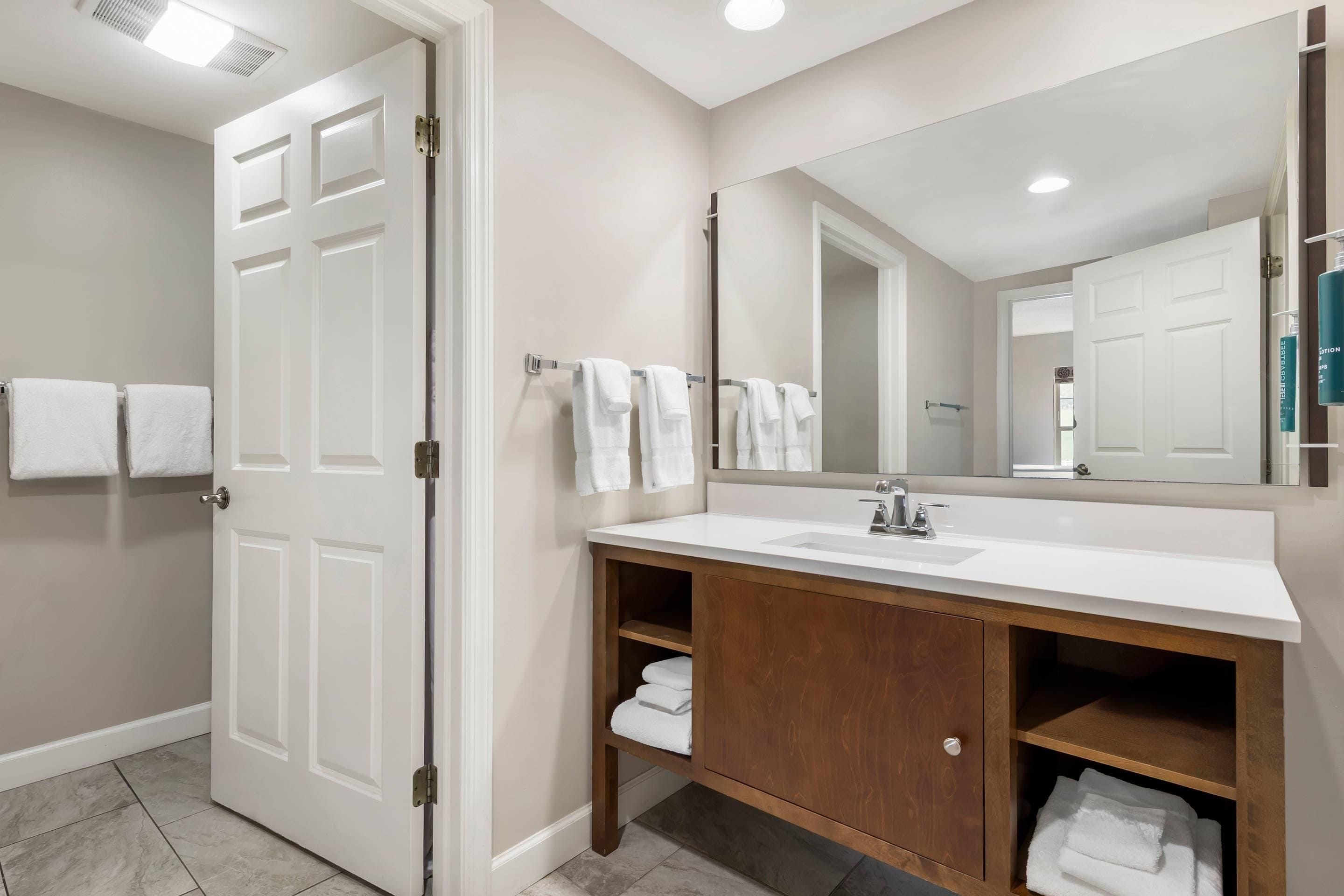 Une salle de bain moderne avec un meuble-lavabo blanc, un grand miroir et une porte donnant sur une autre pièce.