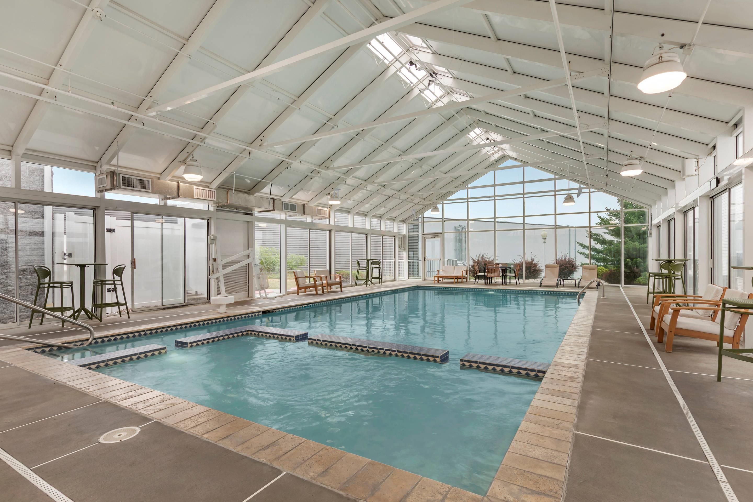 A large indoor swimming pool with multiple lanes, surrounded by lounge chairs and tables, under a high ceiling with skylights.
