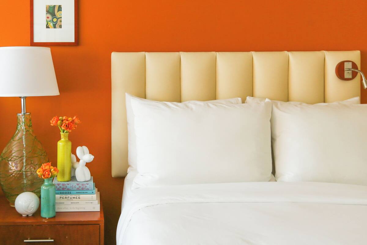 A neatly made bed with white linens, a beige upholstered headboard, and a bedside table with a lamp, a vase with flowers, and a stack of books.
