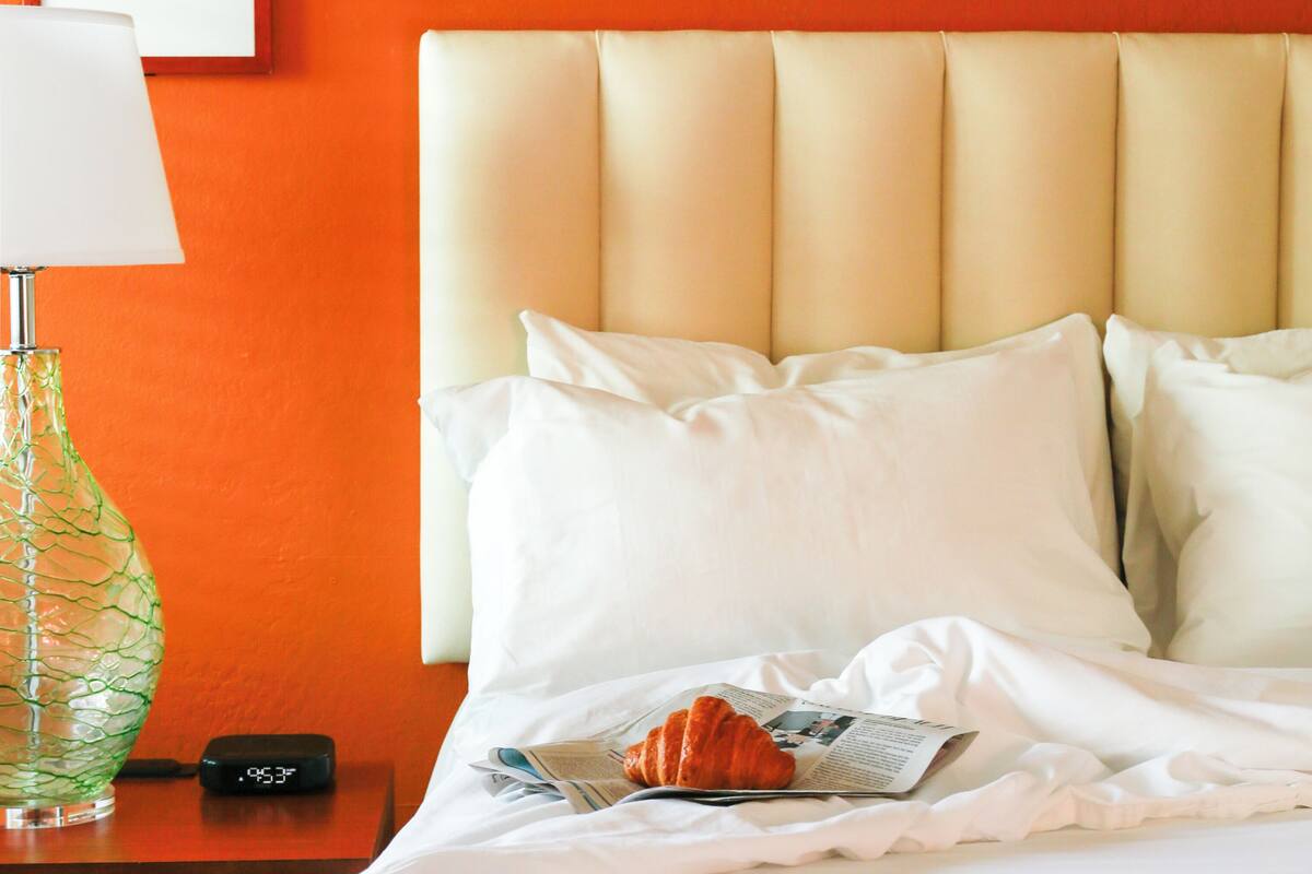A hotel room with a bed, pillows, a lamp, a clock, and a croissant on the bed.