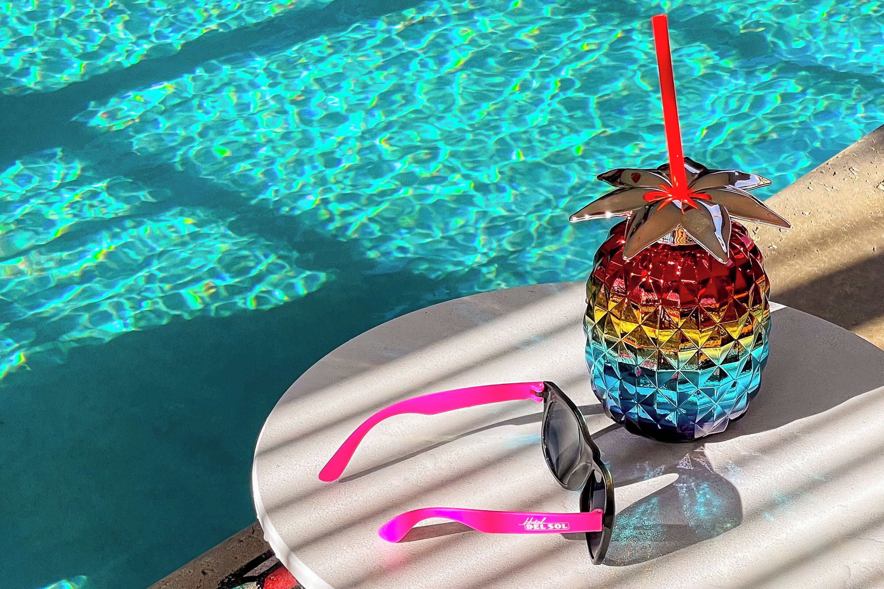 A colorful drink with a straw and sunglasses on a table by a pool.
