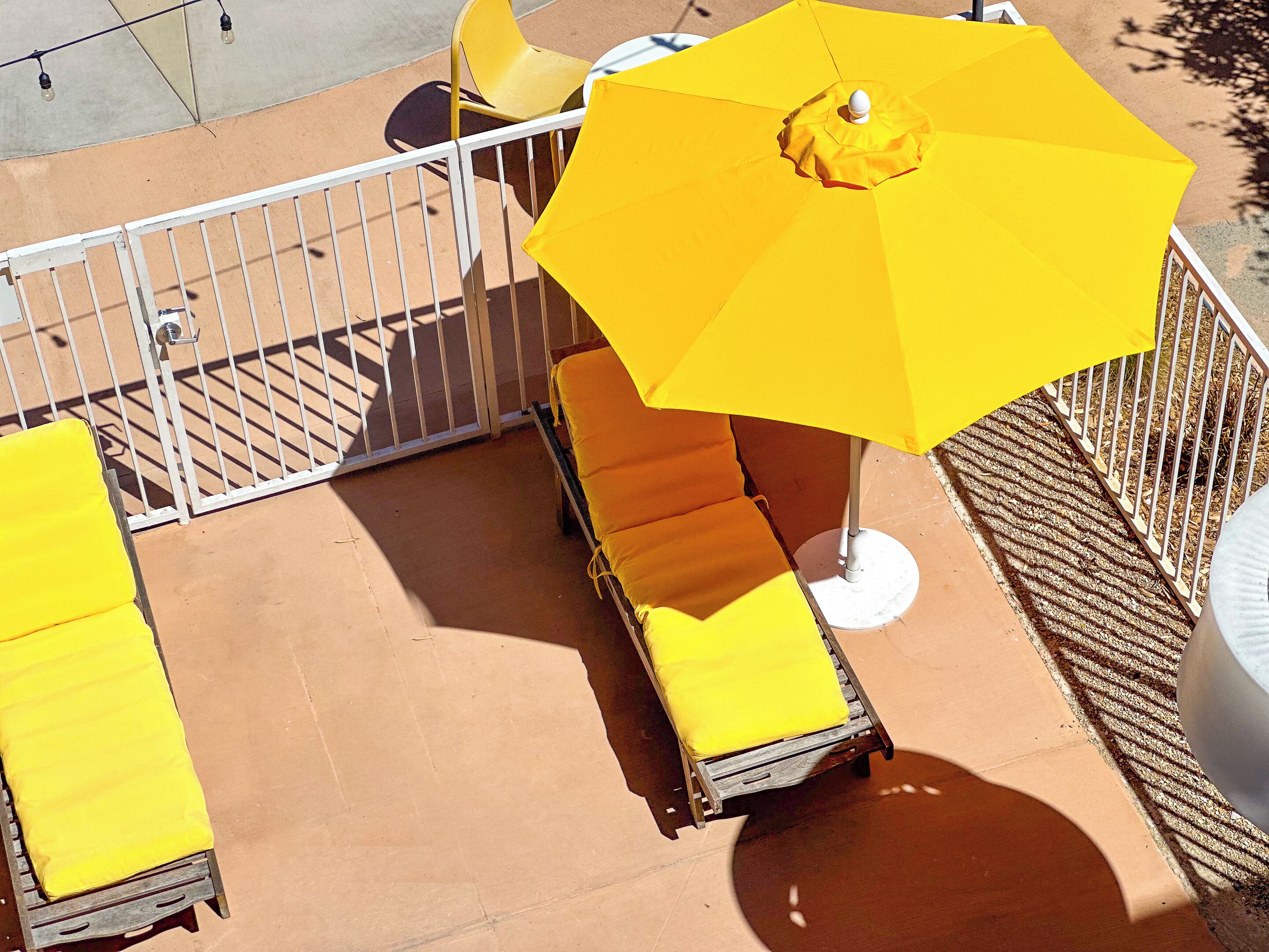 A sunlit outdoor seating area with yellow chairs and an umbrella, enclosed by a white metal fence.