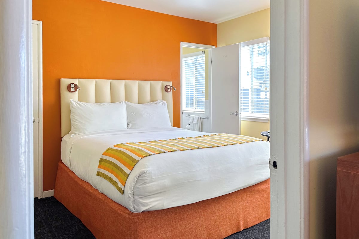 A hotel room with a bed, orange walls, a window with blinds, and a door.
