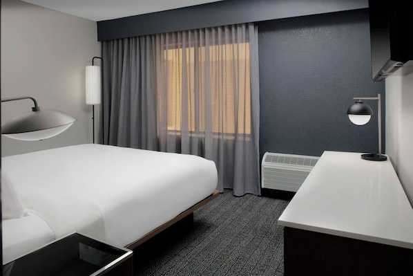 A modern hotel room with a large bed, a desk, a lamp, and a window with curtains.