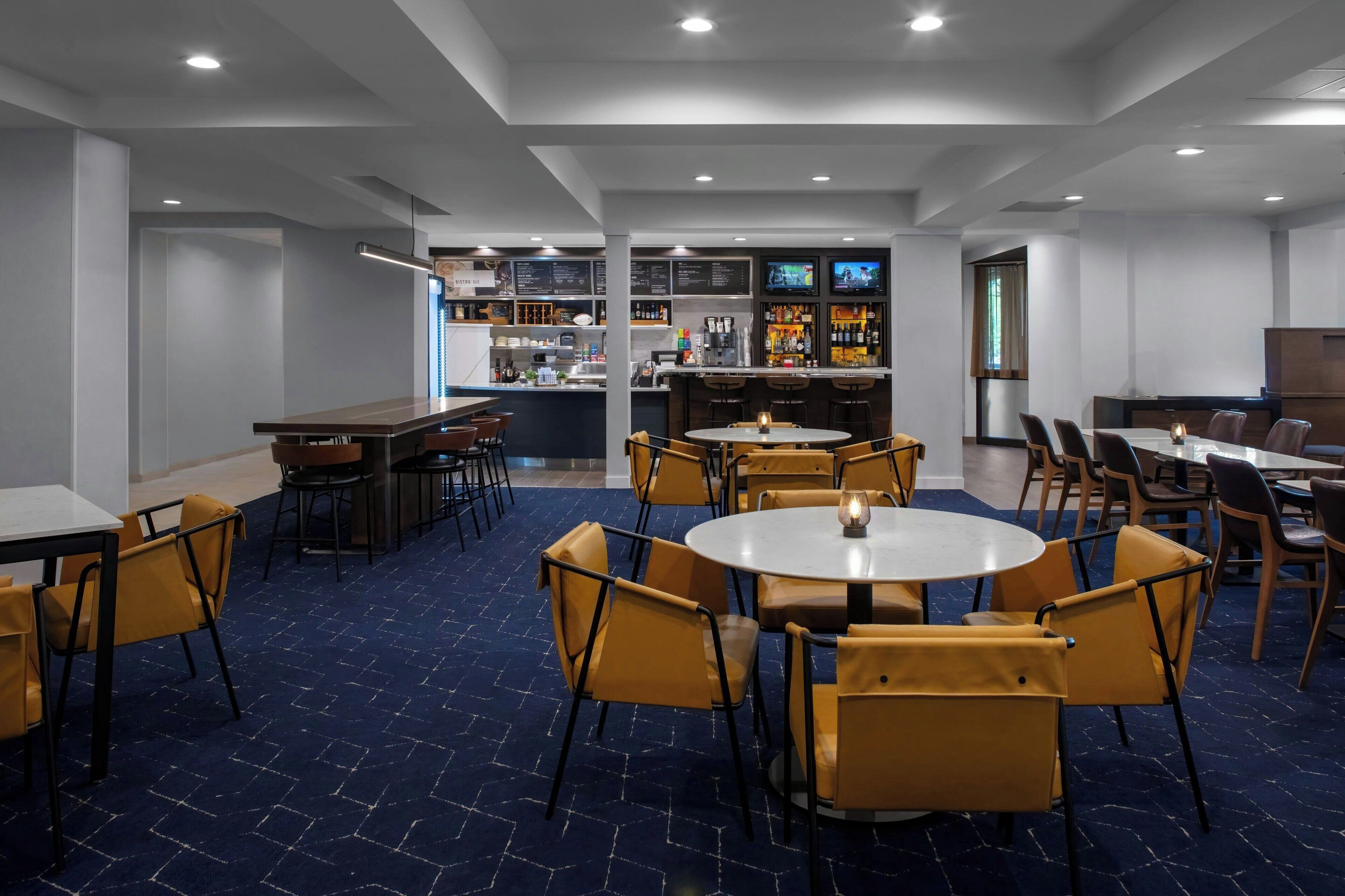 Un espace repas moderne avec des tables rondes et des chaises jaunes, un bar avec un présentoir de menus, et une cuisine en arrière-plan.