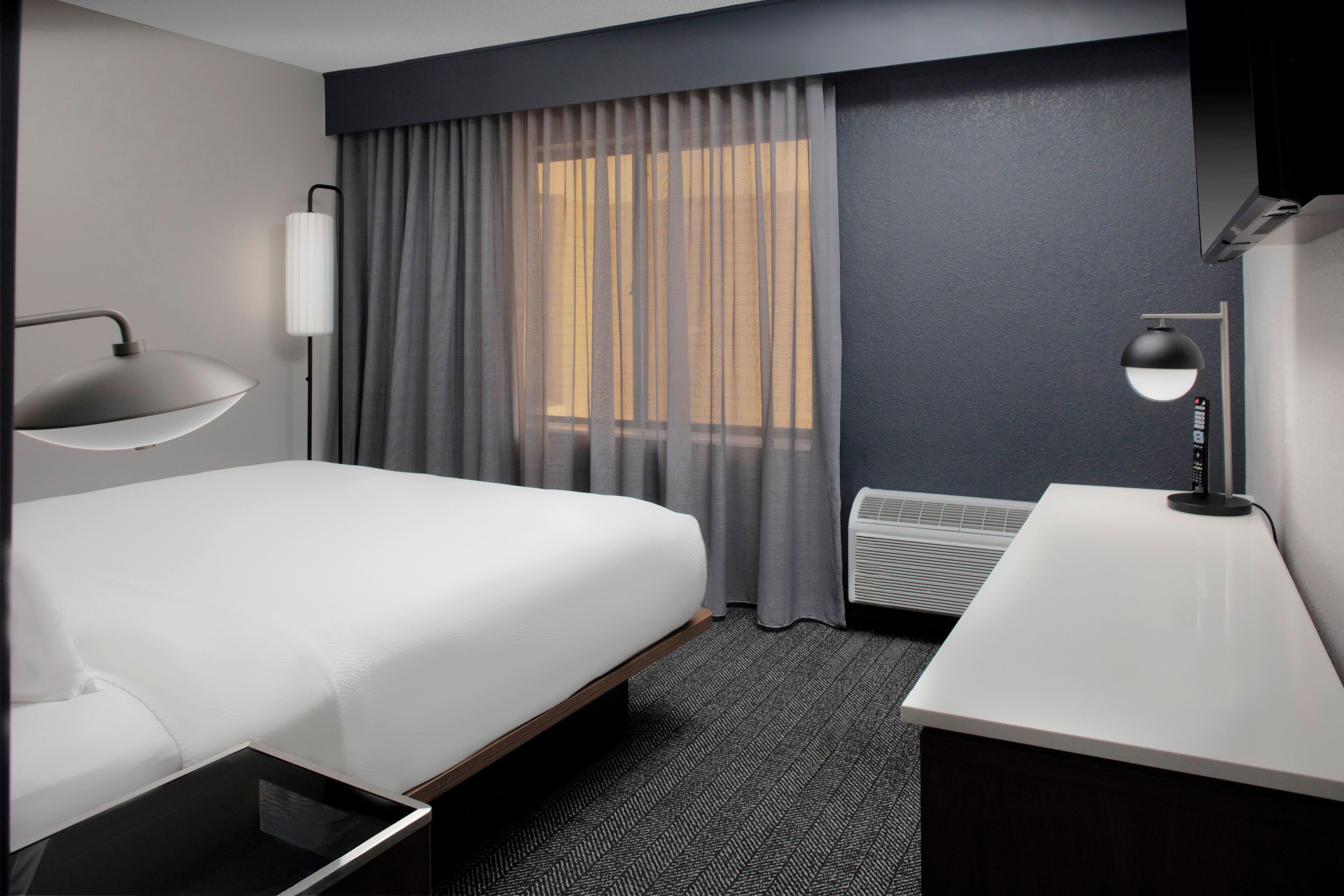 Une chambre d’hôtel moderne avec un grand lit, un bureau, une lampe et une fenêtre avec des rideaux.