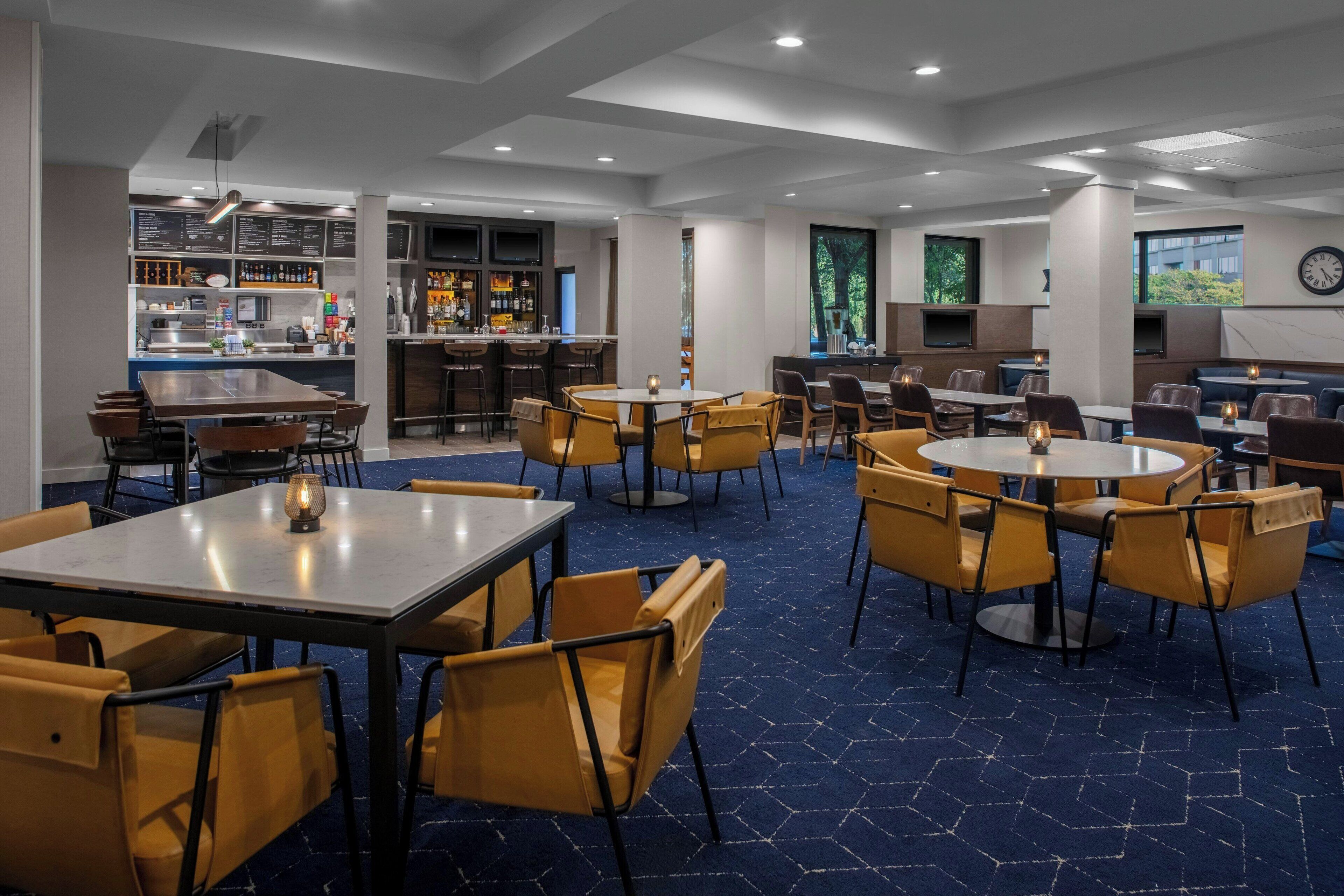 A modern restaurant with multiple seating areas, including round tables and booths, and a bar area with a menu display.