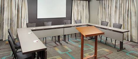 Un salón de conferencias con una larga mesa, sillas, una pantalla de proyector y cortinas.