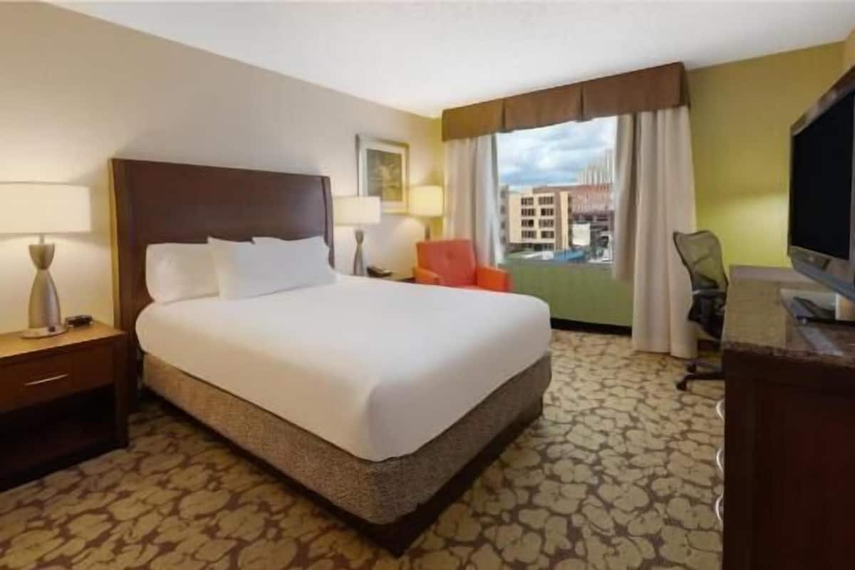 A hotel room with a bed, a desk, a chair, a TV, and a window with curtains.