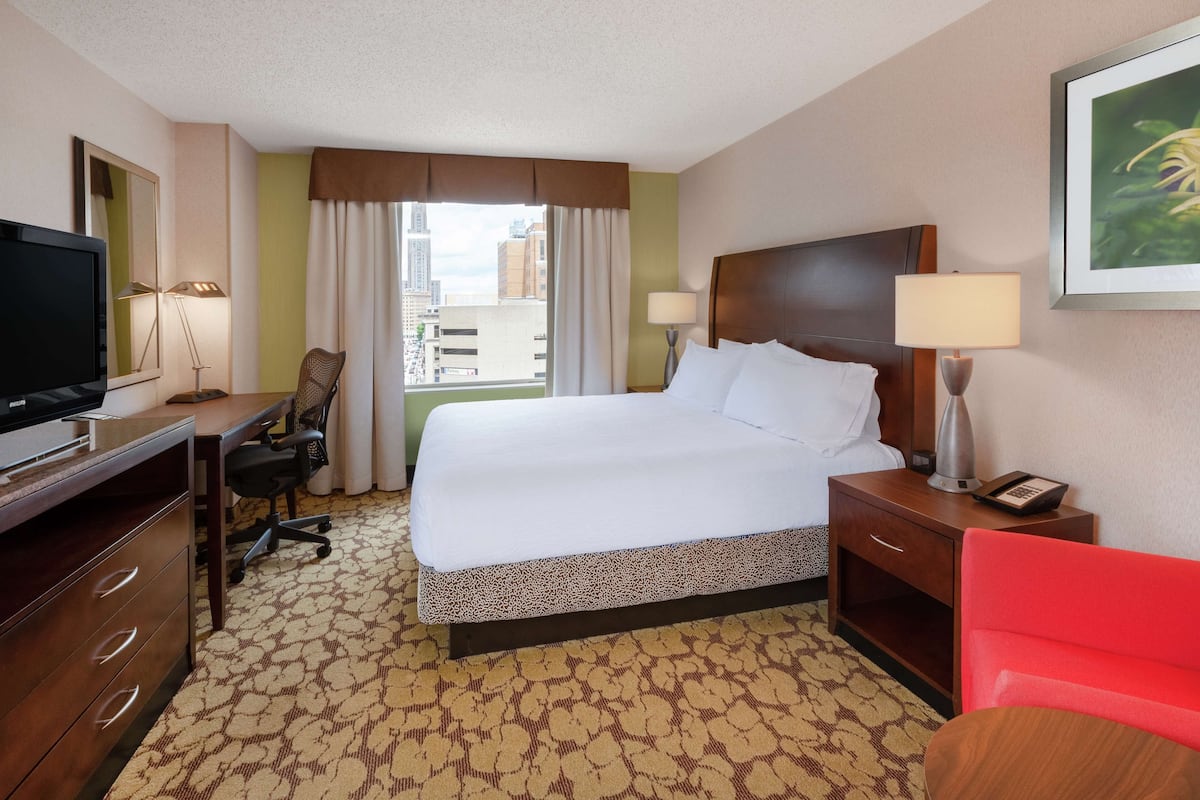 A hotel room with a large bed, a desk with a chair, a television, a nightstand with a lamp, and a window with curtains.