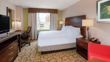 A hotel room with a large bed, a desk with a chair, a television, a nightstand with a lamp, and a window with curtains.
