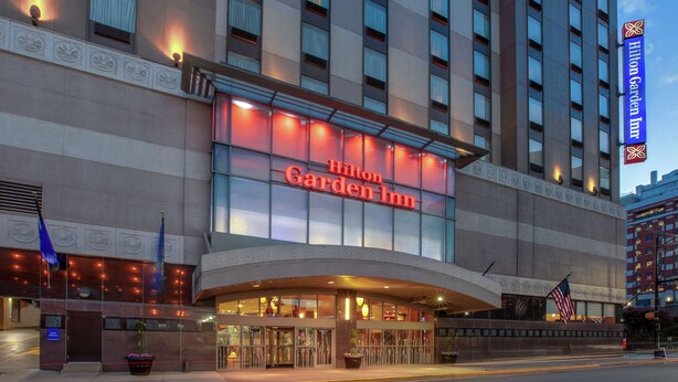L’extérieur de l’hôtel Hilton Garden Inn se distingue par une enseigne lumineuse bien visible, une grande entrée avec des portes vitrées et un drapeau américain exposé à l’extérieur.