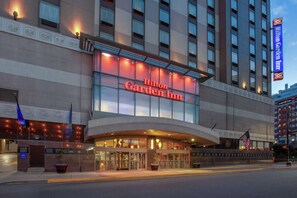 The exterior of the Hilton Garden Inn with a prominent illuminated sign, a large entrance with glass doors, and an American flag displayed outside.