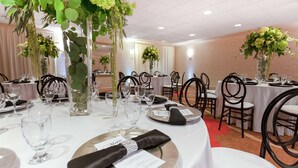 Une salle de banquet avec des tables rondes dressées pour le repas, chacune étant recouverte d’une nappe blanche, entourée de chaises noires et agrémentée de verres transparents.