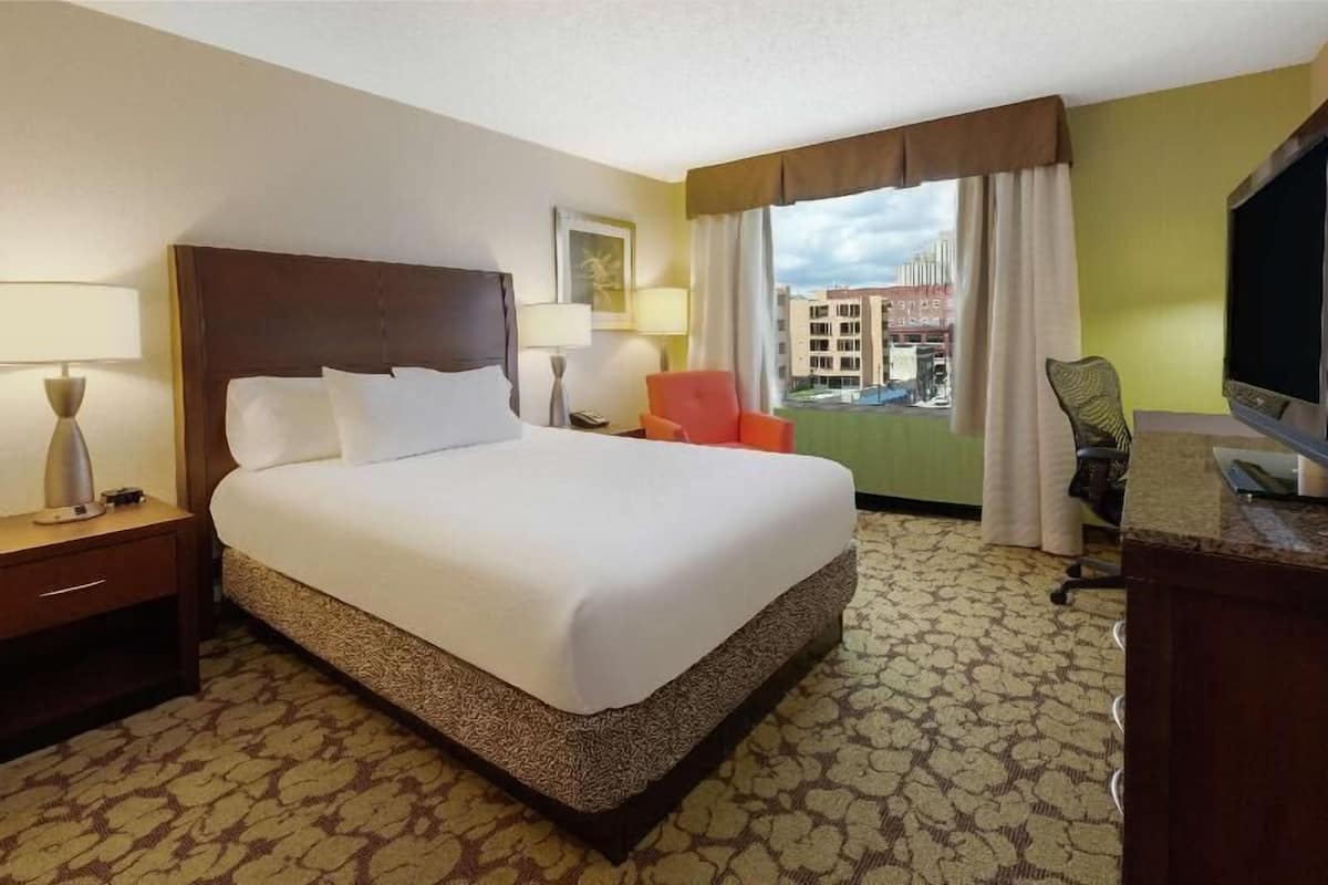 A hotel room with a large bed, a desk, a chair, a TV, and a window with a view of buildings.