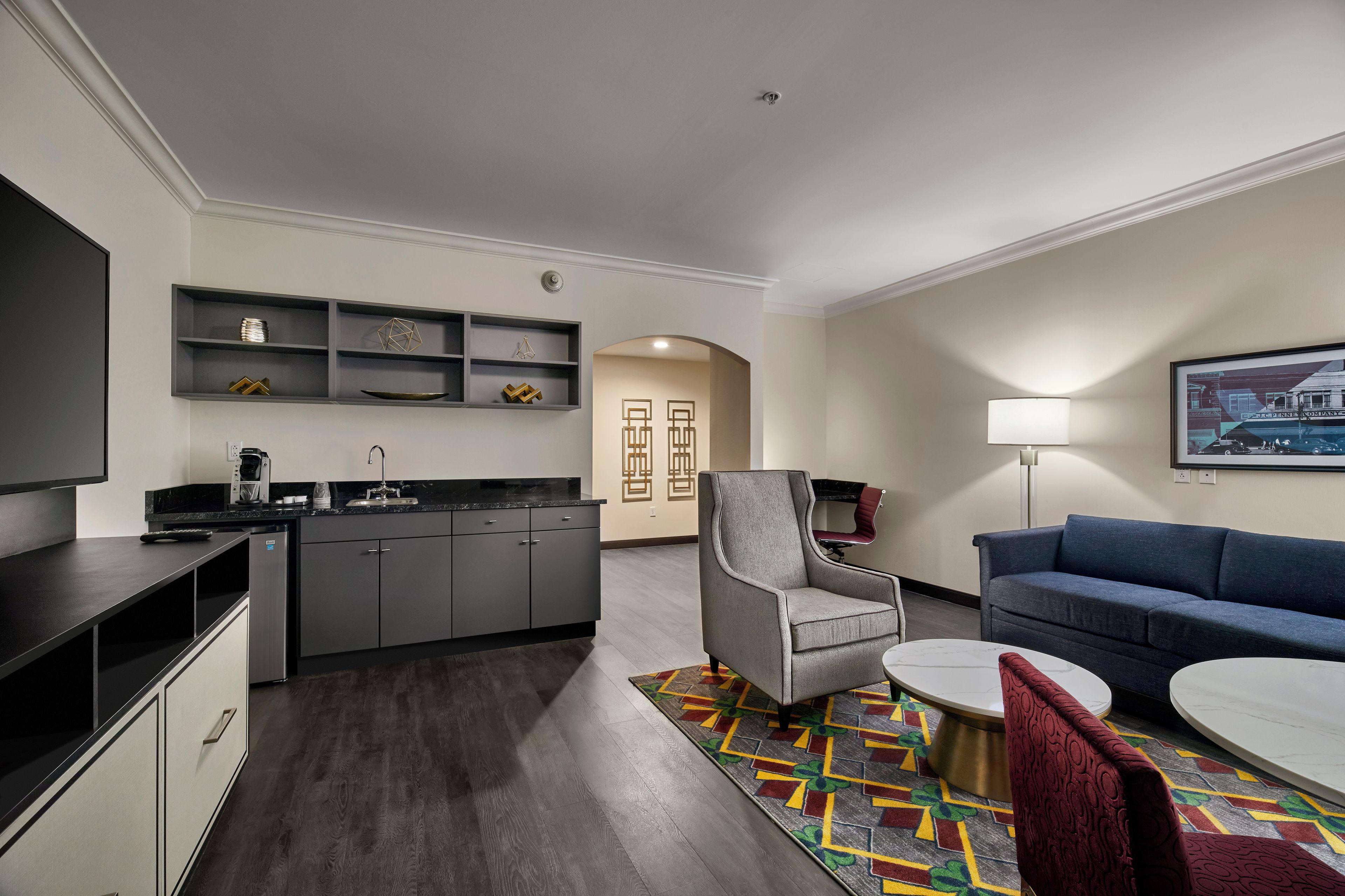 A modern kitchen with dark cabinetry, a built-in coffee machine, and a dining area with a round table and a colorful rug.