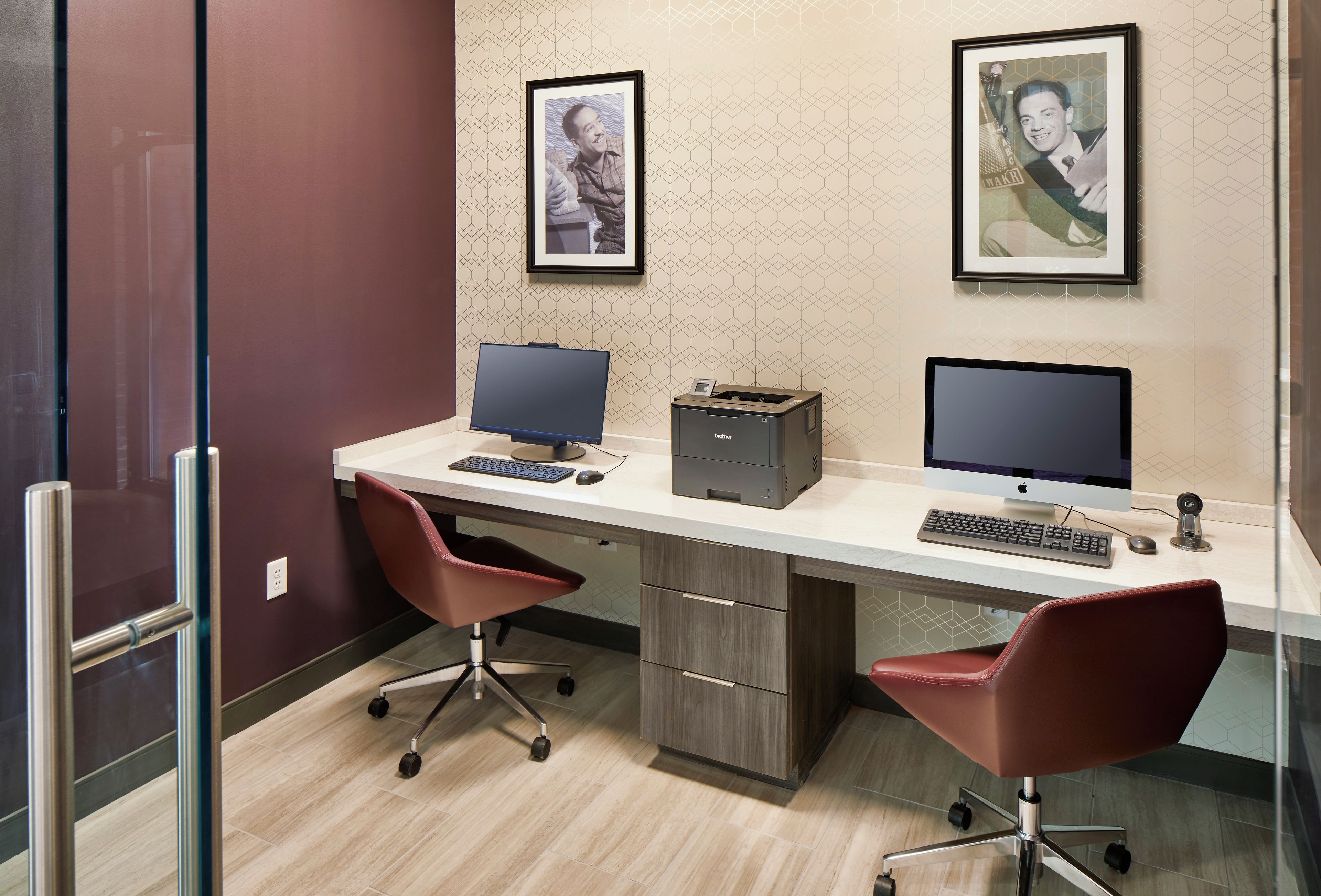 A modern office space with two desks, each with a computer monitor, keyboard, and mouse. There are two red office chairs. The room has a patterned wallpaper and a glass door on the left.