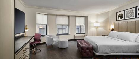 A hotel room with a bed, desk, chair, ottoman, and large windows.