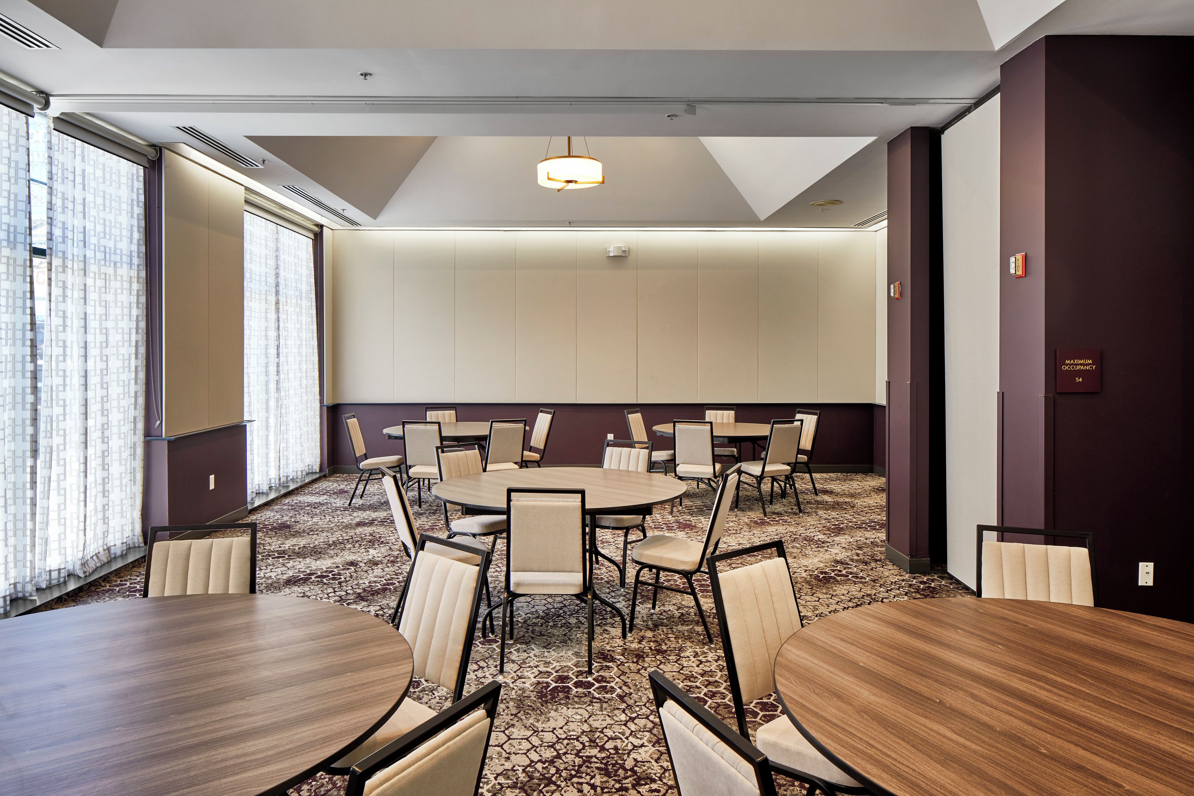 A conference room with multiple tables and chairs, large windows with patterned curtains, and a ceiling with recessed lighting.
