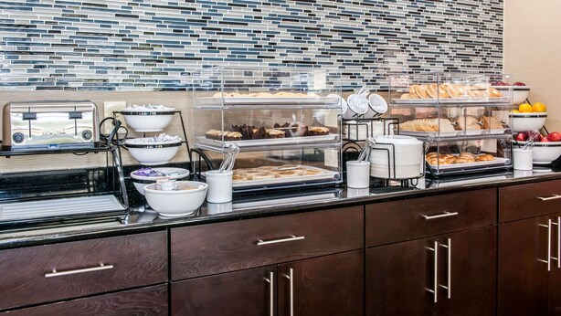 Montaje de bufé con diversos alimentos en recipientes transparentes, una máquina de café y un revestimiento de azulejos.