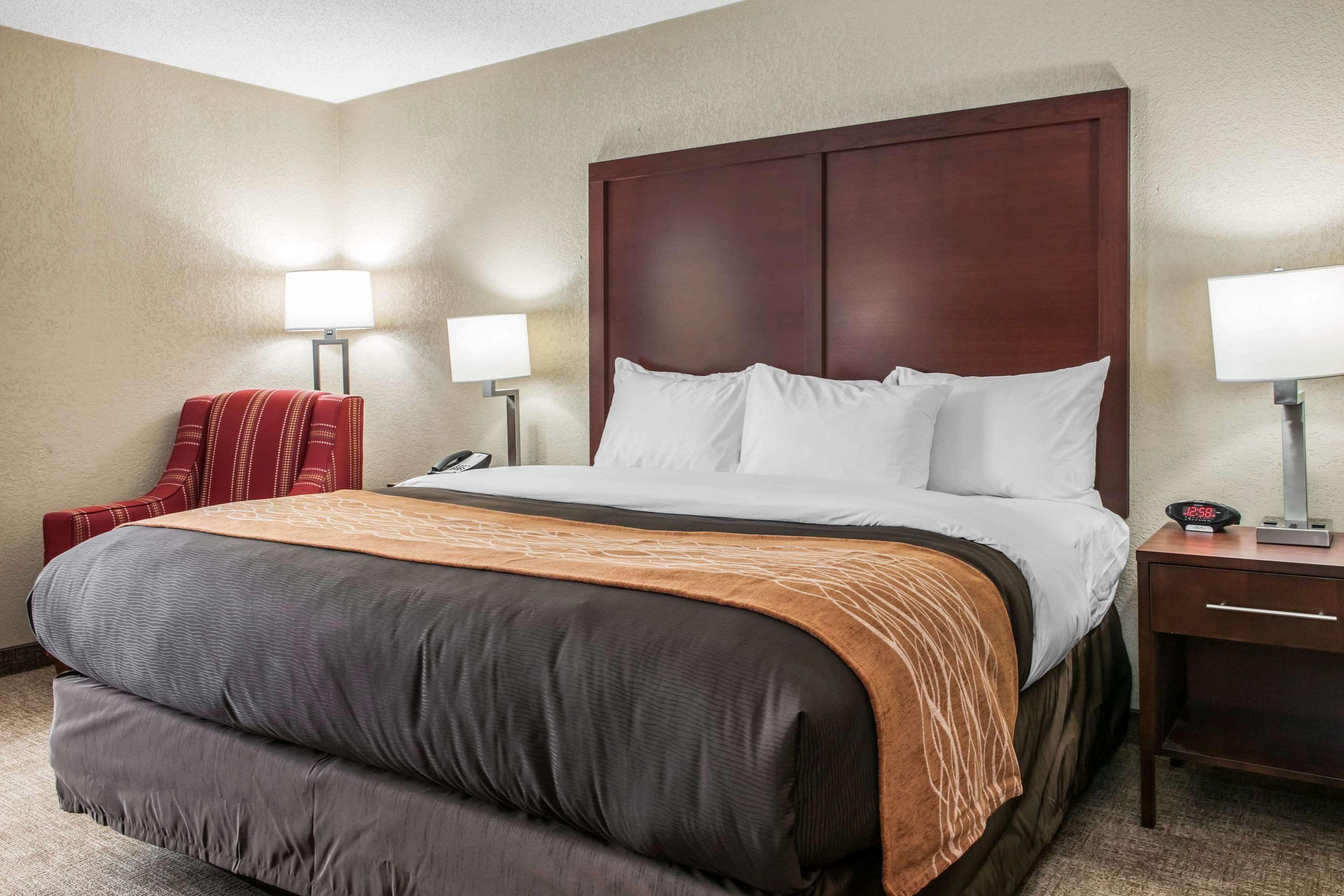 A hotel room with a large bed, two bedside lamps, a red chair, and a nightstand with a clock.