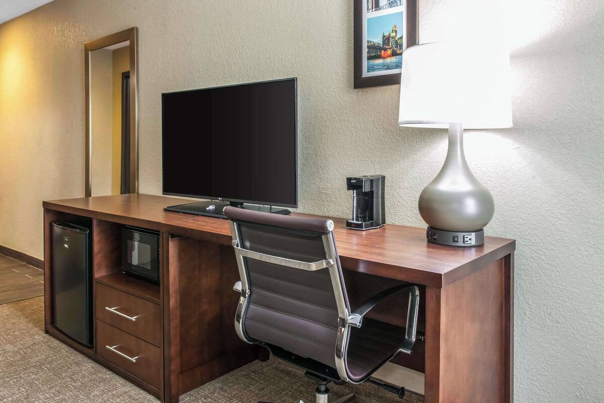 A hotel room with a wooden desk, a flat-screen TV, a lamp, a refrigerator, and a chair.