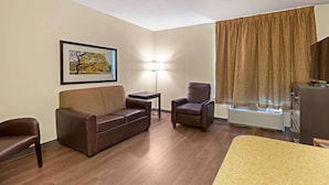 A hotel room with a brown leather sofa, a single armchair, a small table, a standing lamp, and a large window with curtains. - Extended Stay America Suites North Chesterfield Arboretum (Richmond)