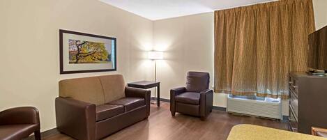 A hotel room with a brown leather sofa, a single armchair, a small table, a standing lamp, and a large window with curtains.