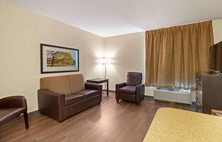Quarto de hotel com sofá de couro marrom, uma poltrona, uma mesinha, abajur e uma grande janela com cortinas.