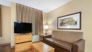 A hotel room with a flat-screen TV, a wooden dresser, a leather sofa, a small table, a lamp, and a framed picture on the wall.