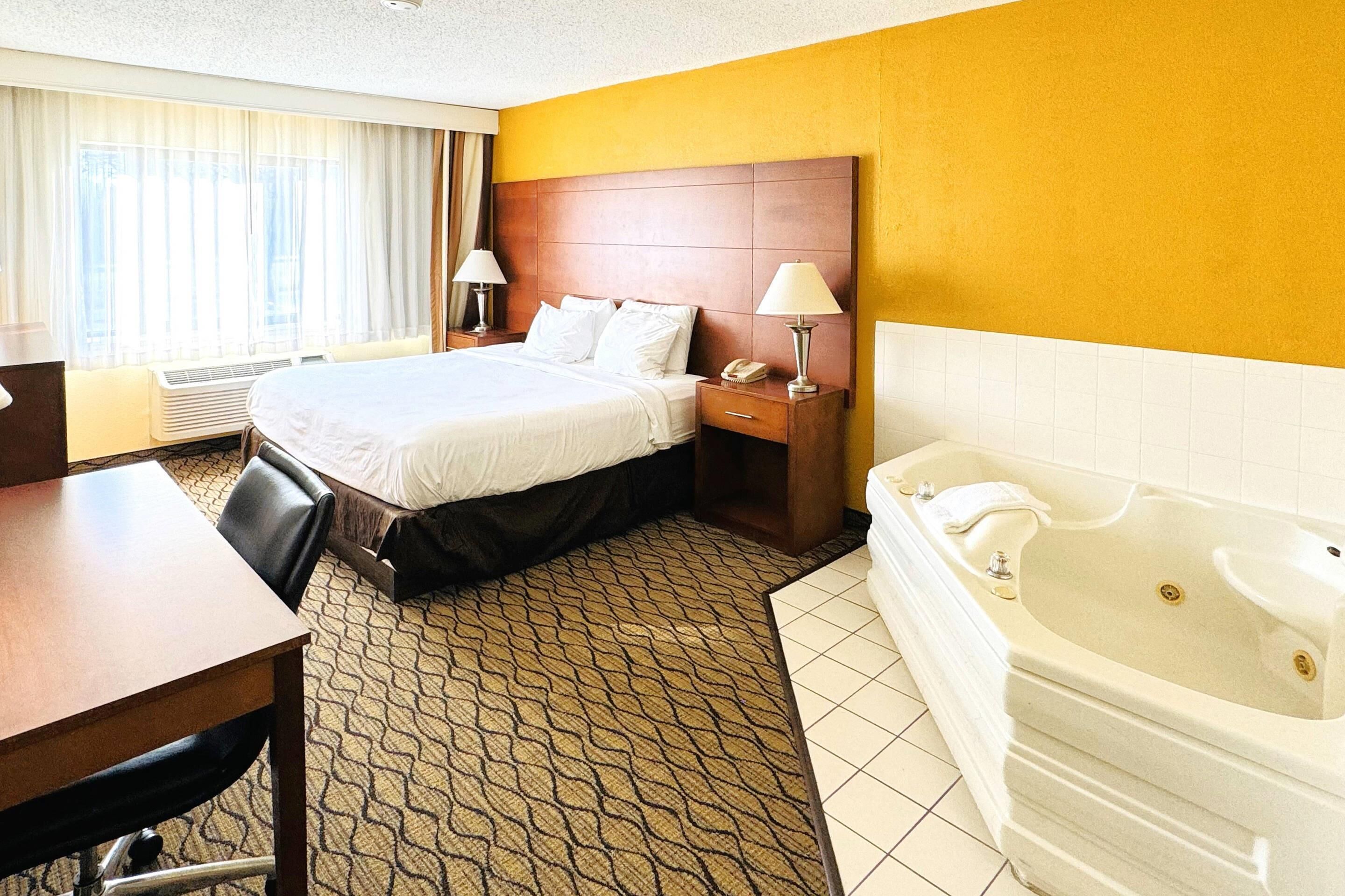 Una habitación de hotel con cama, escritorio, silla, mesita de noche, lámpara y bañera.