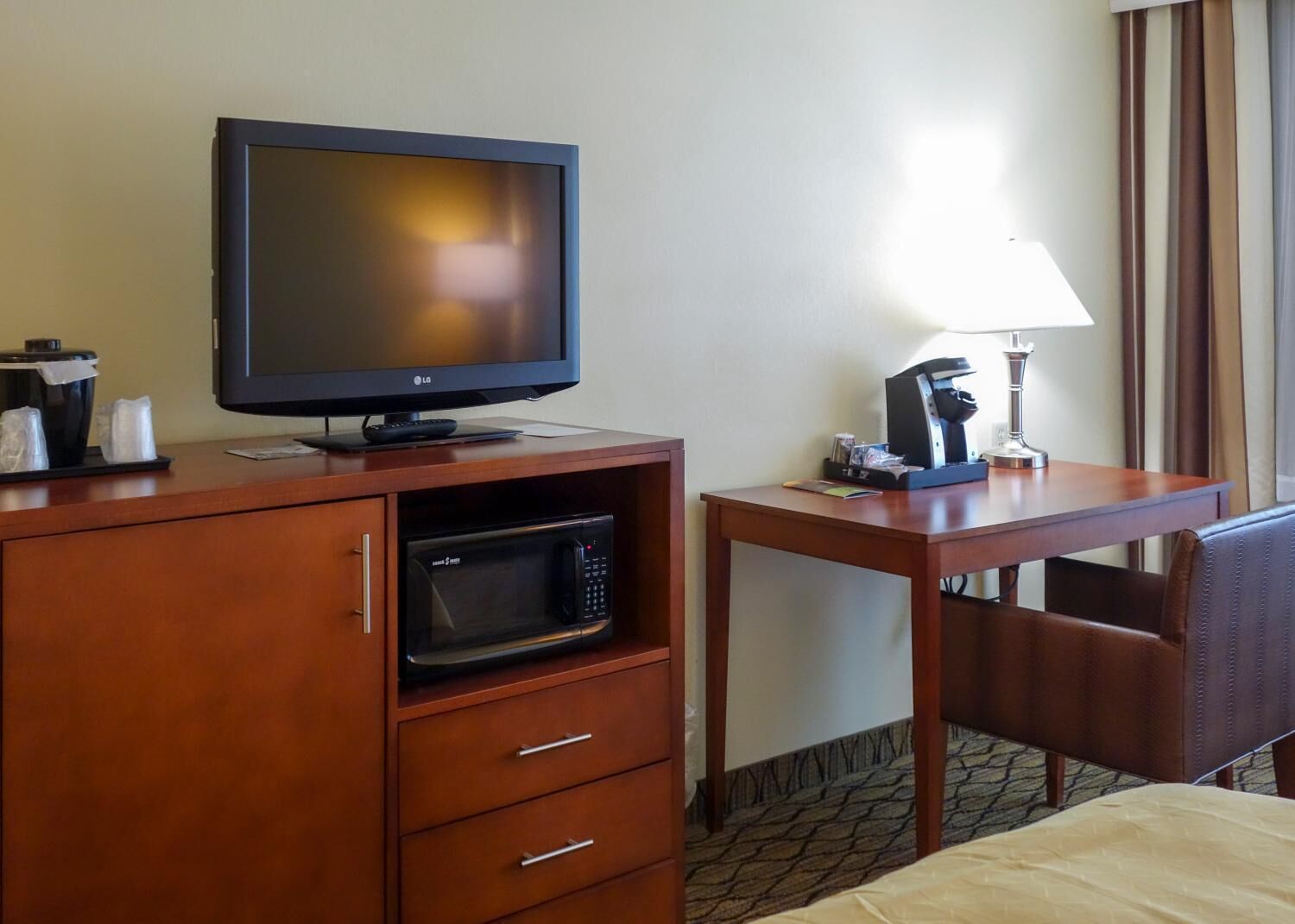Habitación de hotel con escritorio de madera, televisor, microondas, cafetera y una silla.