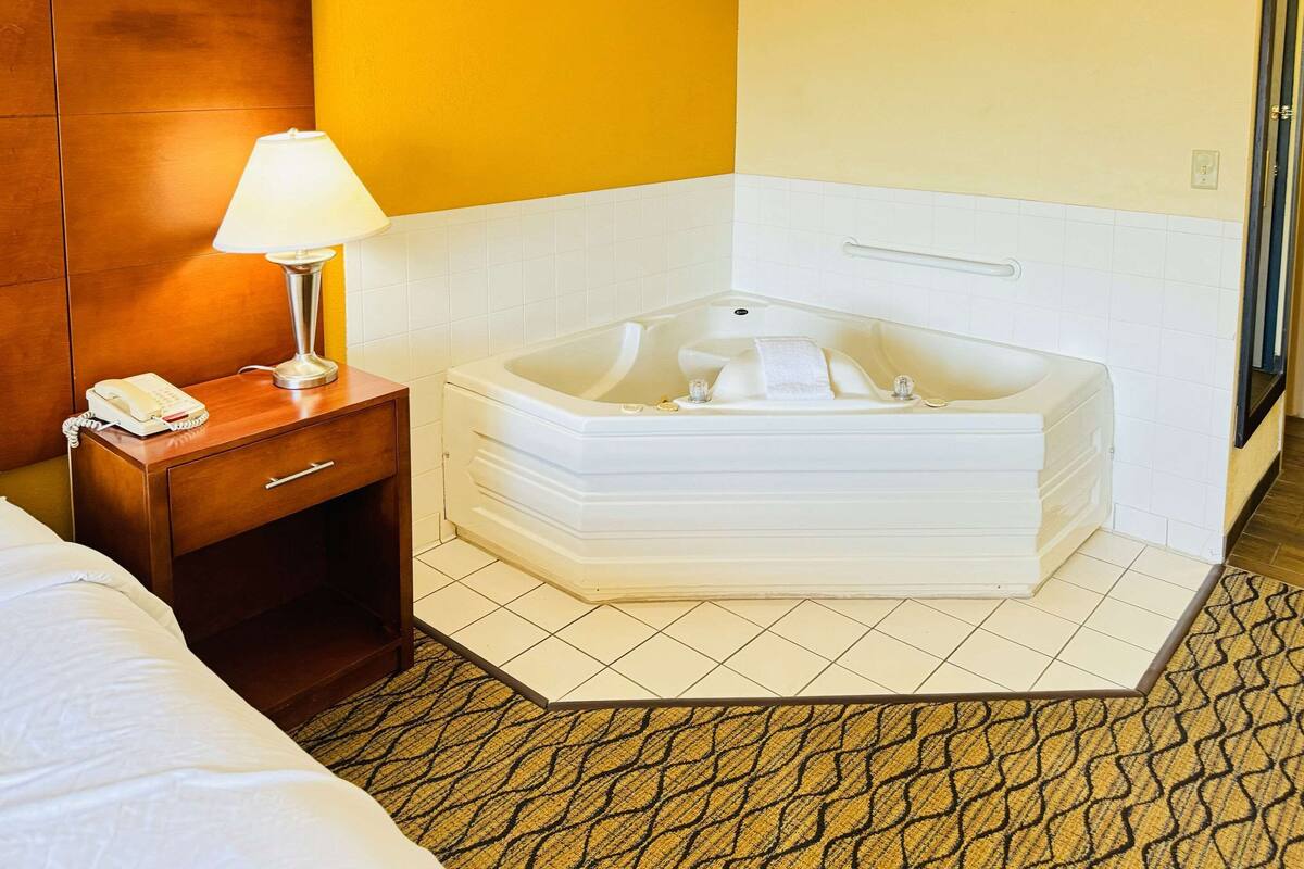 A hotel room with a corner bathtub, a bedside table with a lamp and phone, and a patterned carpet.