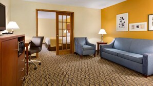 Une chambre d’hôtel comprenant un canapé, deux fauteuils, un bureau avec une lampe, une télévision et une porte donnant sur une autre pièce.
