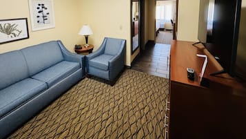 A hotel room with a sofa, armchair, TV, lamp, and artwork on the wall.