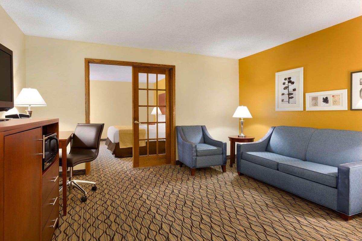 A hotel room with a bed, a desk with a computer, a blue armchair, a grey sofa, and a patterned carpet.
