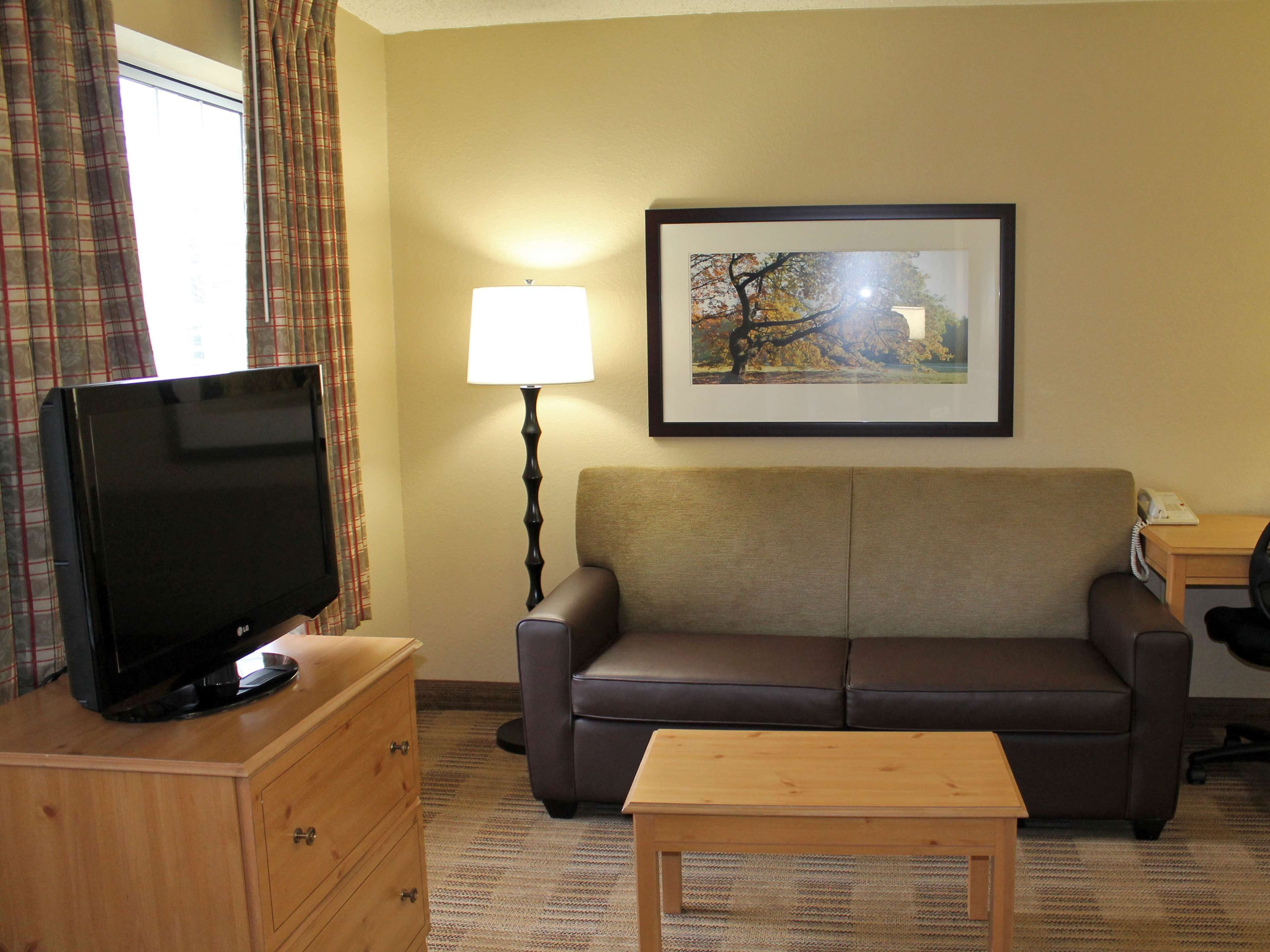 Habitación de hotel con televisión de pantalla plana sobre un tocador de madera, un sofá de cuero marrón, una mesa de centro de madera, una lámpara y un cuadro en la pared.