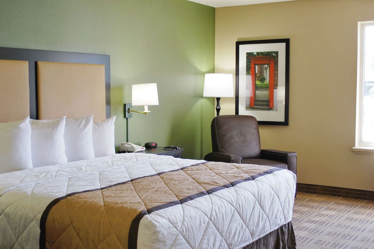 A hotel room with a large bed, two bedside lamps, a chair, a framed picture on the wall, and a window.
