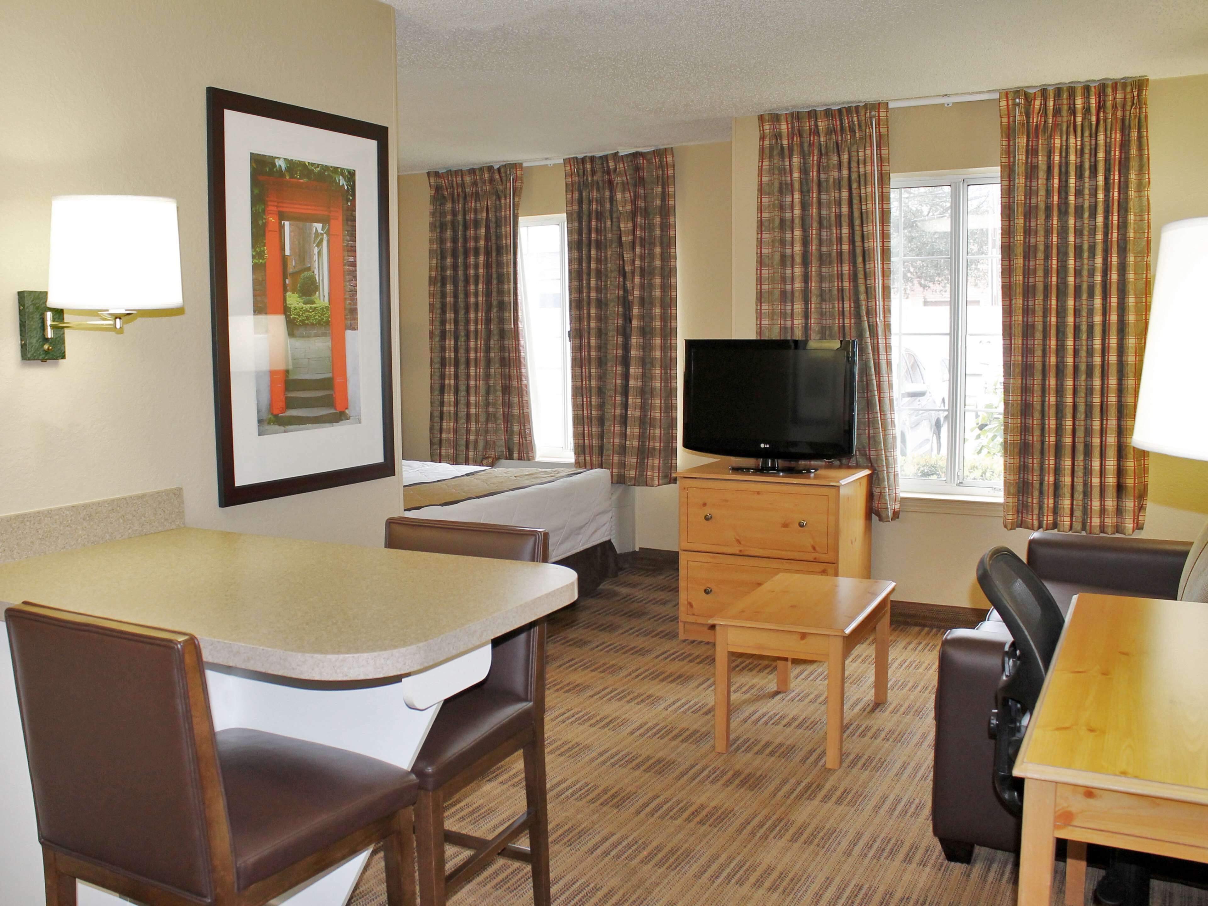 A hotel room with a bed, a TV on a wooden cabinet, a dining area with a table and chairs, and plaid curtains on the windows.