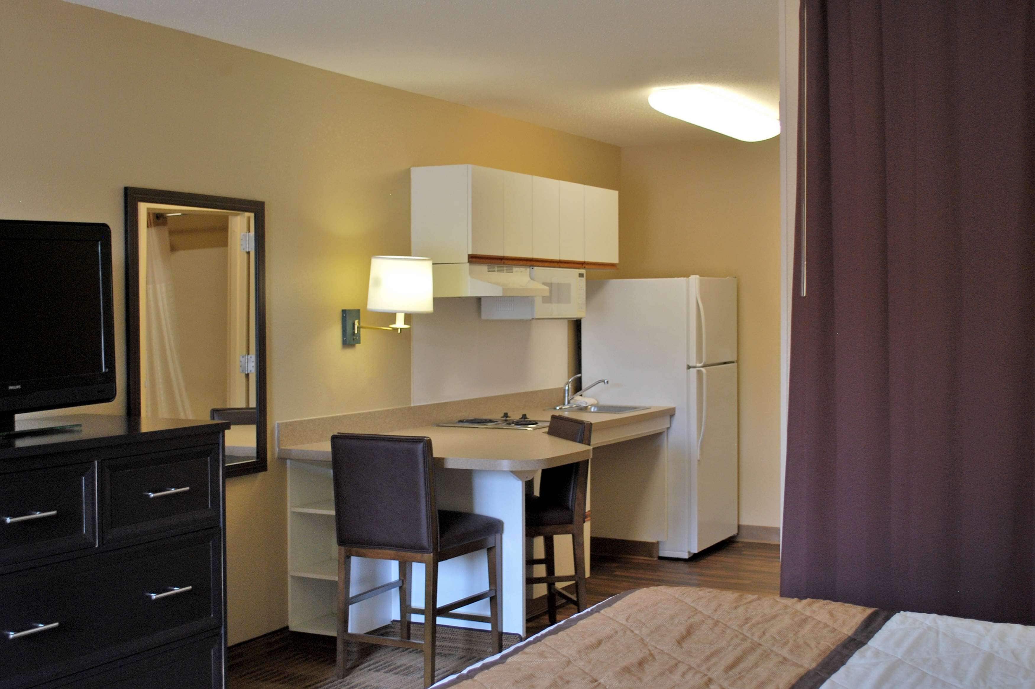 Habitación de hotel compacta con kitchenette, un televisor de pantalla plana, un escritorio con dos sillas y una cama.
