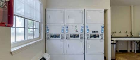 A laundry room with six stacked washing machines, a window with blinds, a radiator, and a table with a trash can.