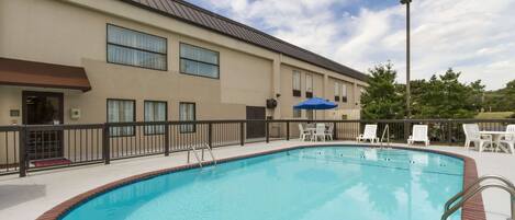 A swimming pool with a brick border, surrounded by a fence and white lounge chairs.