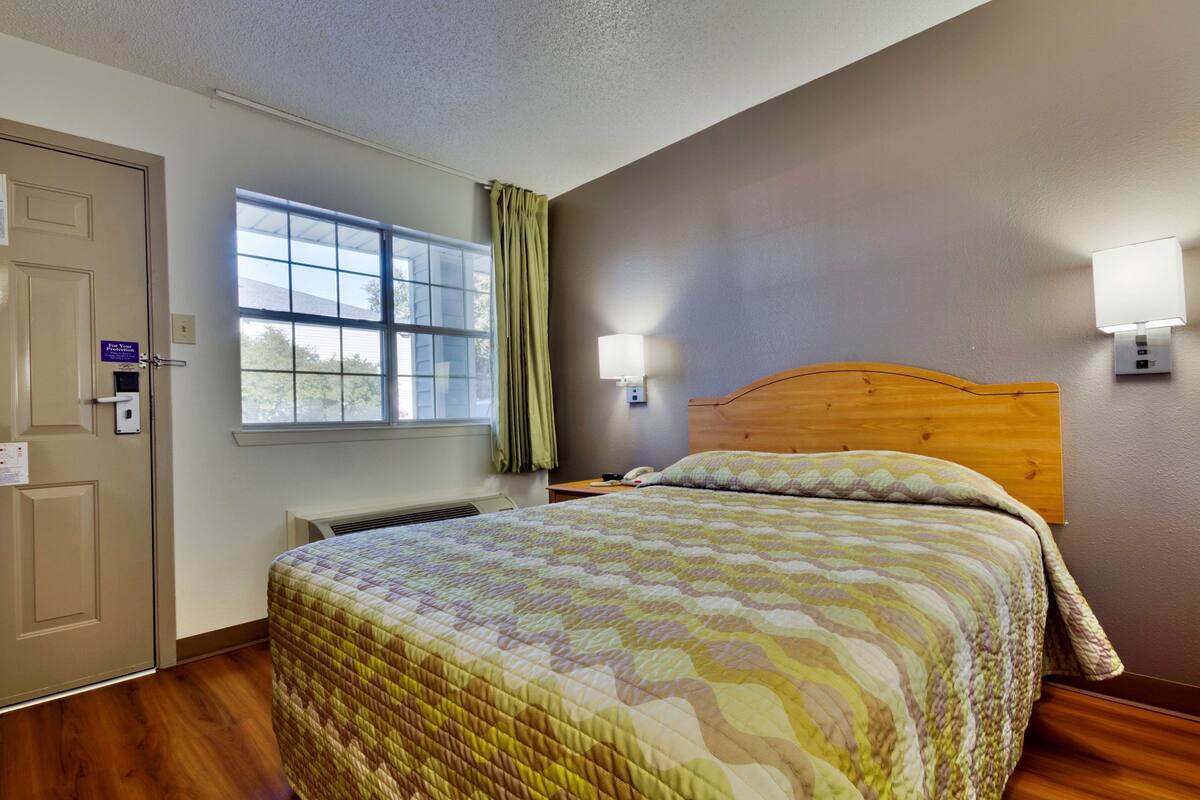 A hotel room with a wooden headboard, a large bed with a patterned quilt, a window with curtains, a wall-mounted lamp, and a thermostat on the wall.
